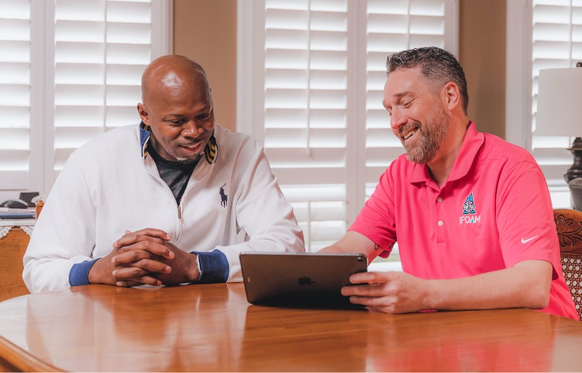 An iFOAM attic insulation contractor sitting at a dining room table with a customer reviewing project details on a tablet